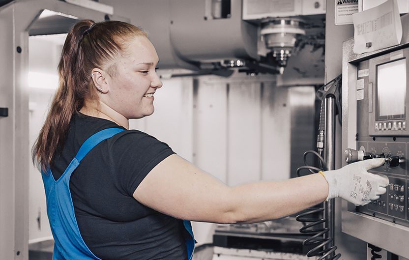 Mitarbeiterin in blauer Arbeitskleidung arbeitet an einer Maschine in einer Fabrikhalle.