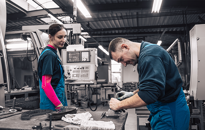 Zwei Mitarbeiter in blauer Arbeitskleidung arbeiten in einer Produktionsumgebung neben einer Maschine an einem Tisch.