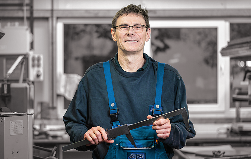 Mitarbeiter in blauer Arbeitskleidung mit Brille hält ein Werkzeug in der Hand und lächelt.