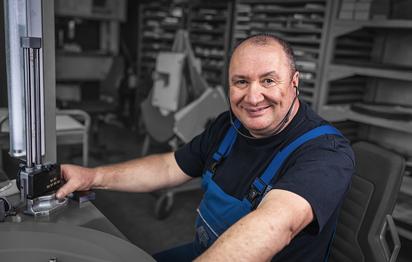 Mitarbeiter in blauer Arbeitskleidung sitzt an einer Maschine und blickt in die Kamera.